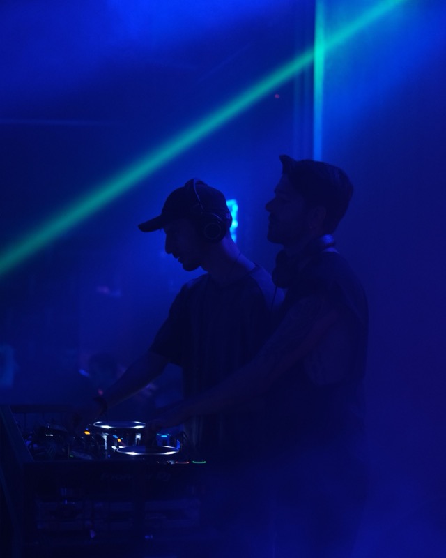 Two DJs at the decks under blue and green club light.